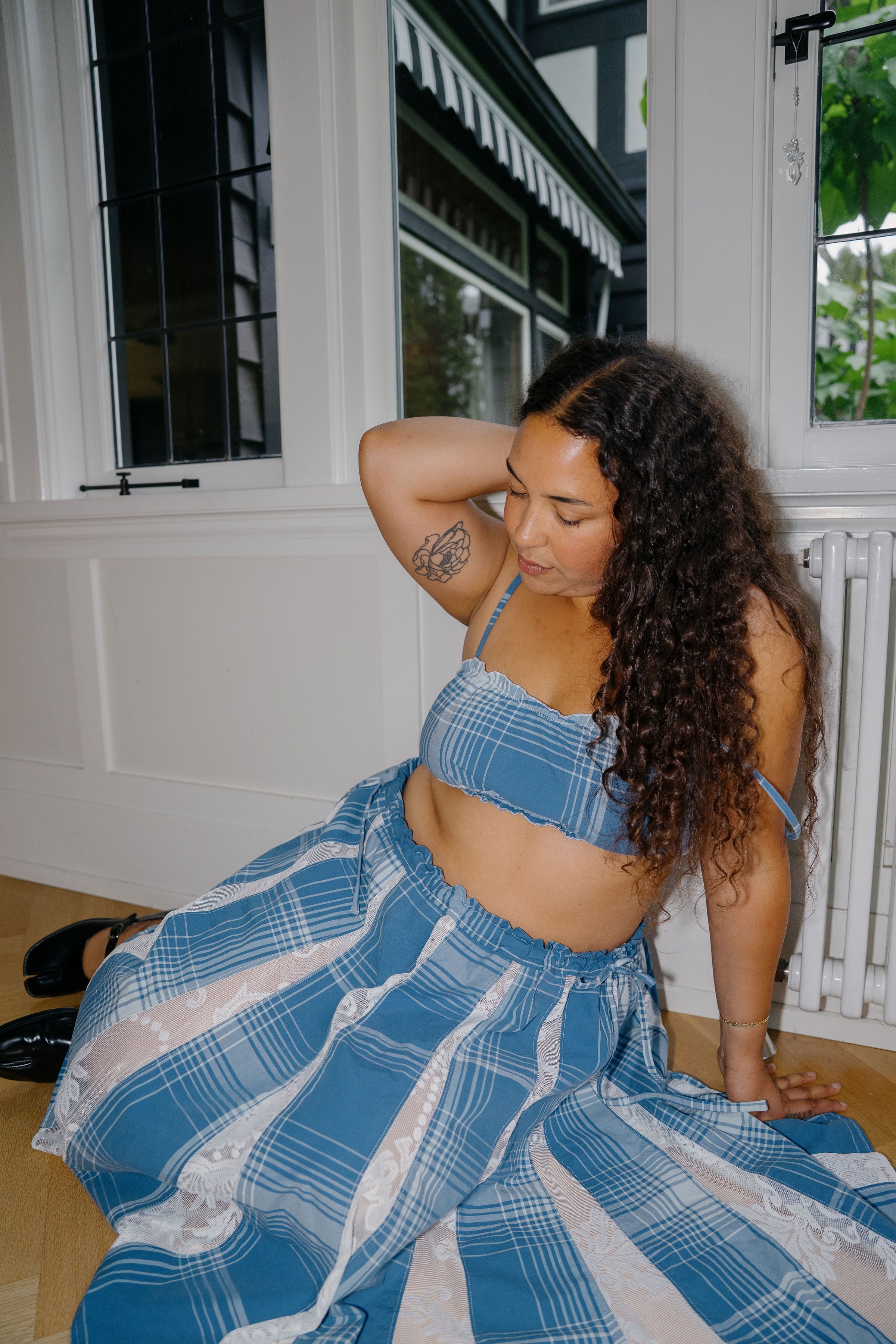A woman with long curly dark hair sits on a wooden floor indoors, wearing a blue and white plaid two-piece outfit consisting of a cropped tank top and a matching wide skirt.